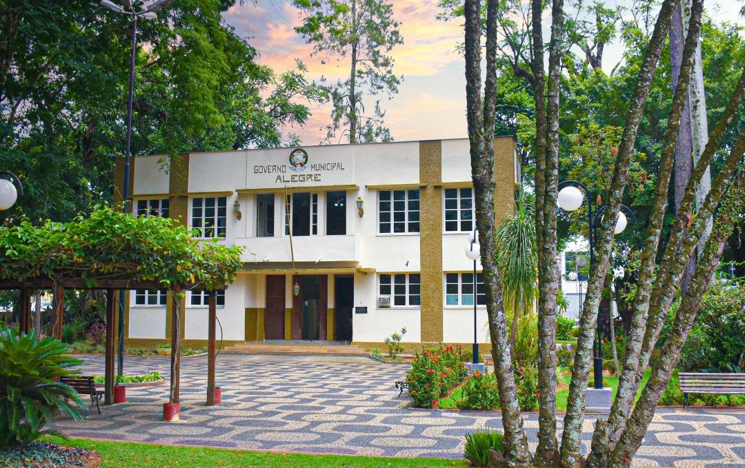 Fachada da sede da Prefeitura de Alegre, com prédio administrativo ao fundo, área arborizada, calçamento em padrão ondulado e bancos na praça em frente.
