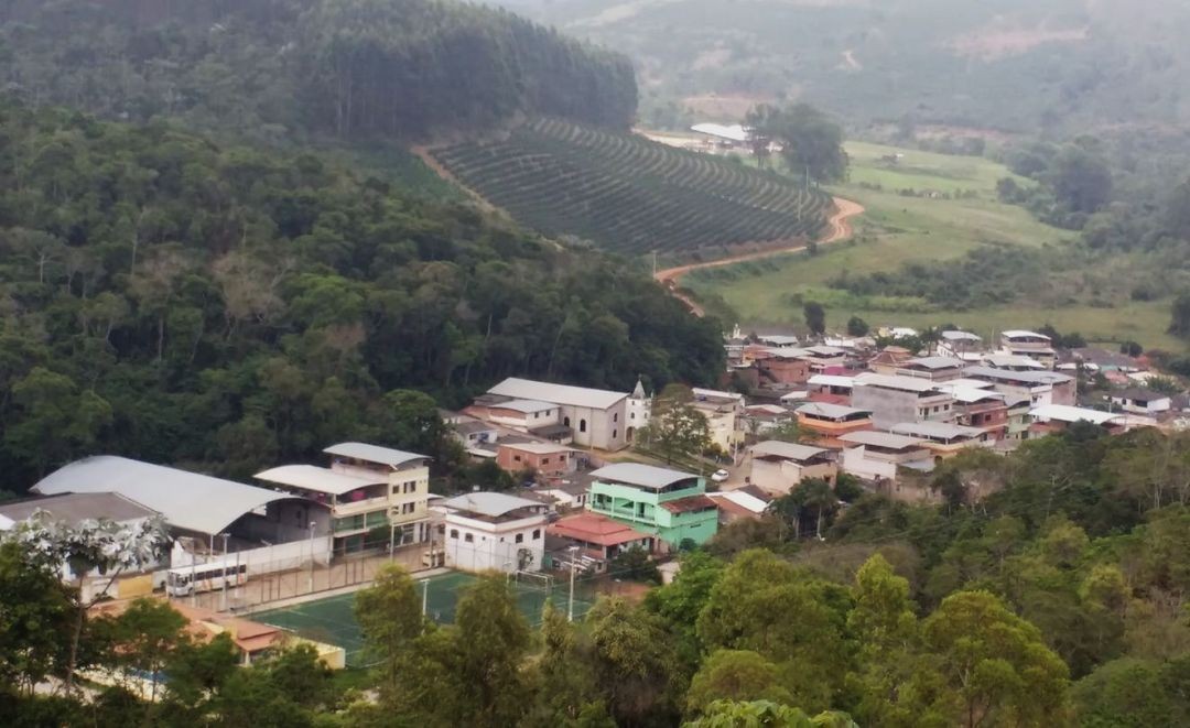 Vista aérea da comunidade de Santa Cruz, em Irupi, com casas, igreja, quadra esportiva e áreas rurais ao redor, incluindo plantações e vegetação.
