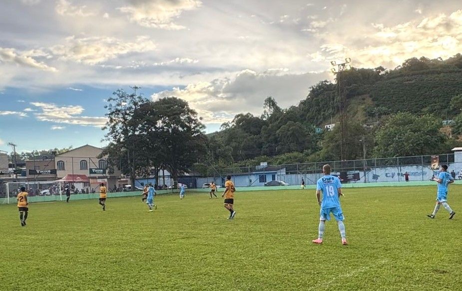 Jogadores disputam partida de futebol do campeonato regional, em campo gramado no Estádio Municipal de Dores do Rio Preto, com equipes uniformizadas de azul e outra de amarelo e preto, tendo ao fundo um alambrado, onde estão algumas pessoas assistindo e casas da cidade, sob um céu amarelado pelo entardecer.