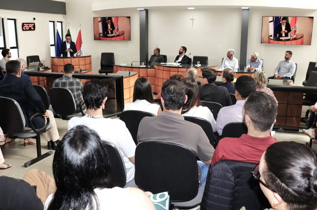 Vista da plenária da Câmara Municipal de Irupi. No fundo, autoridades compõem a mesa diretora e telões laterais transmitem o discurso do prefeito de Irupi em exercício Abercílio Machado. Em primeiro plano, o público composto por servidores e gestores assiste à cerimônia de costas para a câmera.
