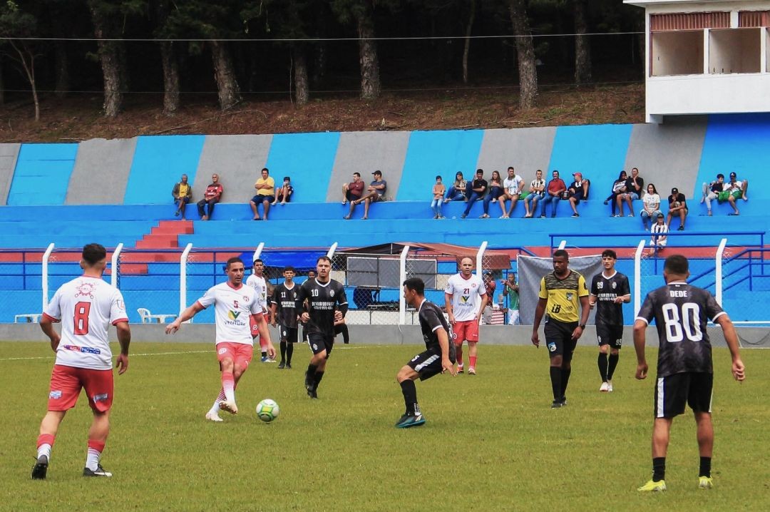 Fotografia de uma partida de futebol em andamento num campo de grama verde. Jogadores vestem uniformes nas cores branco e preto. Ao fundo, vê-se a arquibancada do estádio com listras verticais em azul e cinza, onde torcedores acompanham sentados.