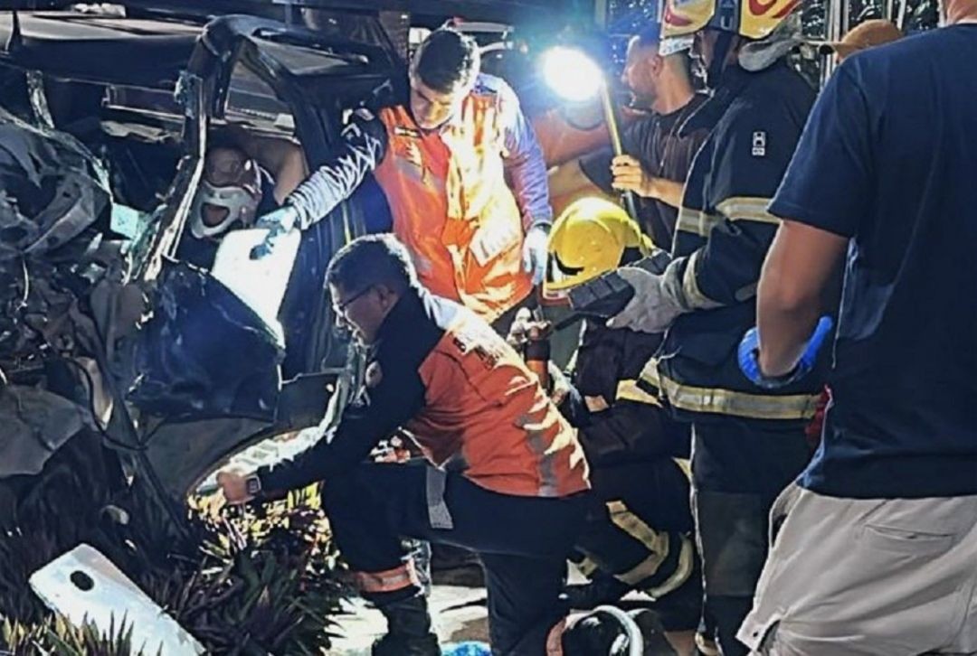 Fotografia noturna mostrando o trabalho das equipes de resgate. Profissionais do SAMU, com uniformes laranjas, e bombeiros, com equipamentos de proteção individual, trabalham juntos para retirar uma vítima de dentro de um veículo prata severamente retorcido. É possível ver a vítima usando um colar cervical enquanto recebe atendimento.