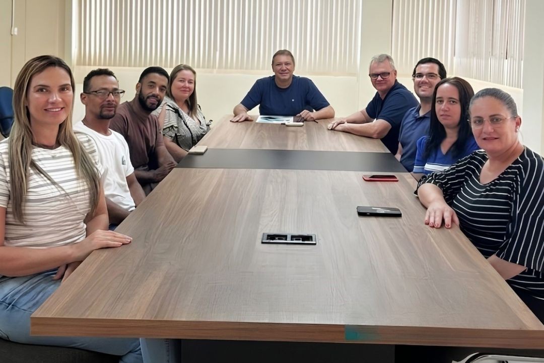Foto em plano médio de uma reunião em torno de uma grande mesa de madeira clara. No centro, ao fundo, o presidente do MEPES, Darci Schaefer, e o prefeito Reginaldo lideram a conversa. Nas laterais da mesa, estão sentadas as vereadoras Adriana e Mychely, além de representantes da EFA e da Associação de Pais. O ambiente é iluminado, com janelas ao fundo cobertas por persianas claras.