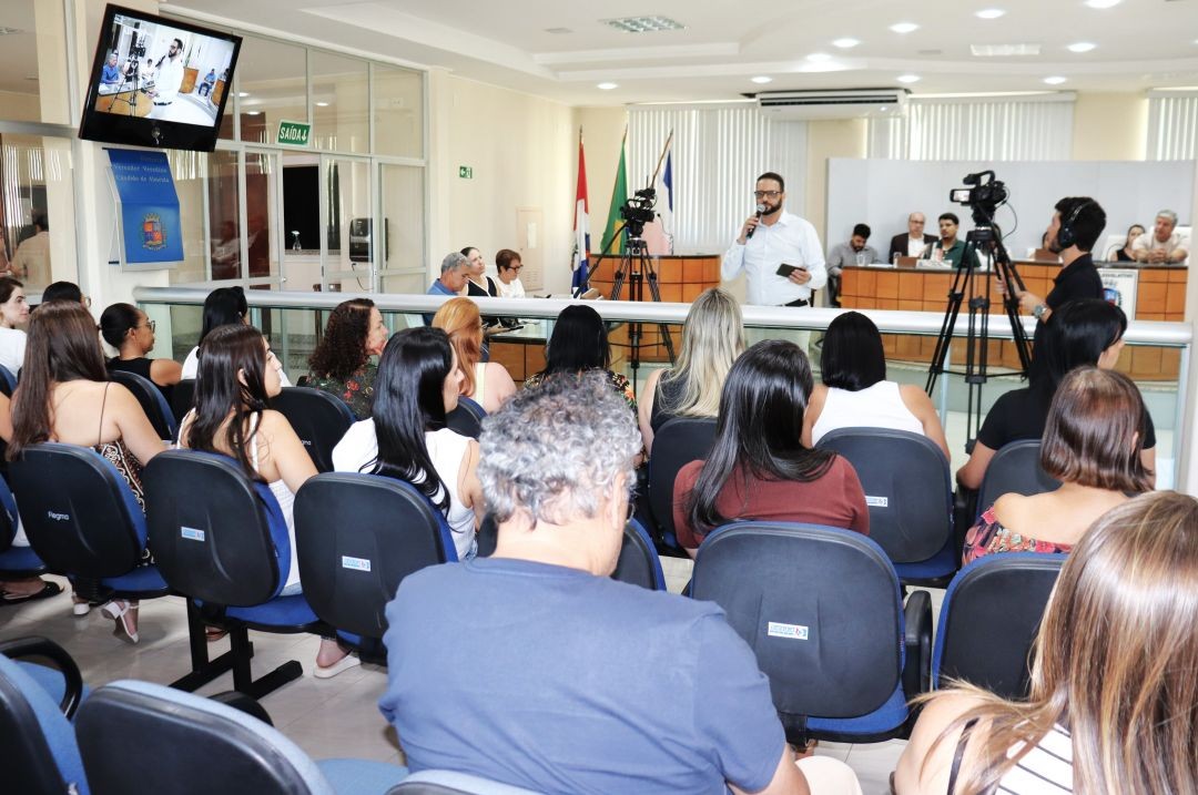Vista do plenário da Câmara Municipal de Iúna durante a audiência de saúde. Em primeiro plano, o público sentado em cadeiras azuis acompanha a apresentação. Ao fundo, o secretário de saúde Waldren Marcelo fala ao microfone, com câmeras de transmissão posicionadas à sua frente.