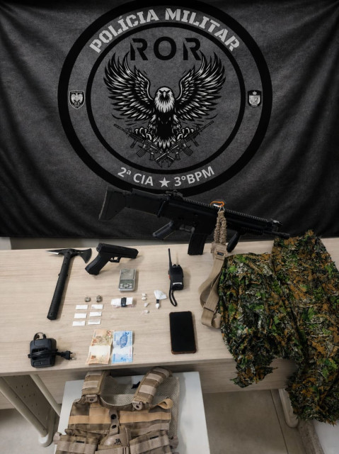Materiais apreendidos pela Polícia Militar em Dores do Rio Preto, incluindo simulacros de armas, drogas, dinheiro, balança de precisão e equipamentos táticos sobre uma mesa.