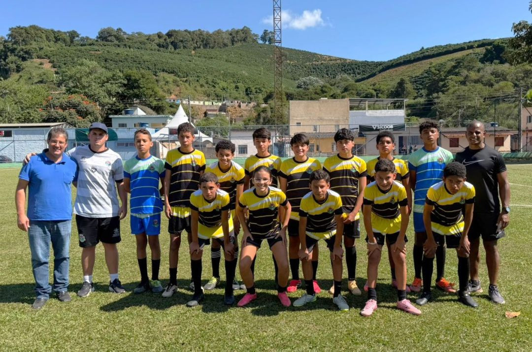 Fotografia de um time de futebol juvenil masculino e feminino posando em um campo de grama natural sob luz solar. Os atletas vestem uniformes listrados em preto e amarelo ou azul e verde. Três adultos (comissão técnica) acompanham a equipe. Ao fundo, uma paisagem montanhosa com plantações de café e céu limpo.