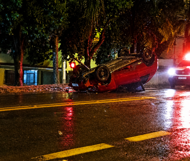 Carro capotado com as rodas para cima na BR-482, em Guaçuí, após colisão com semáforo; pista molhada e equipes de atendimento no local à noite.