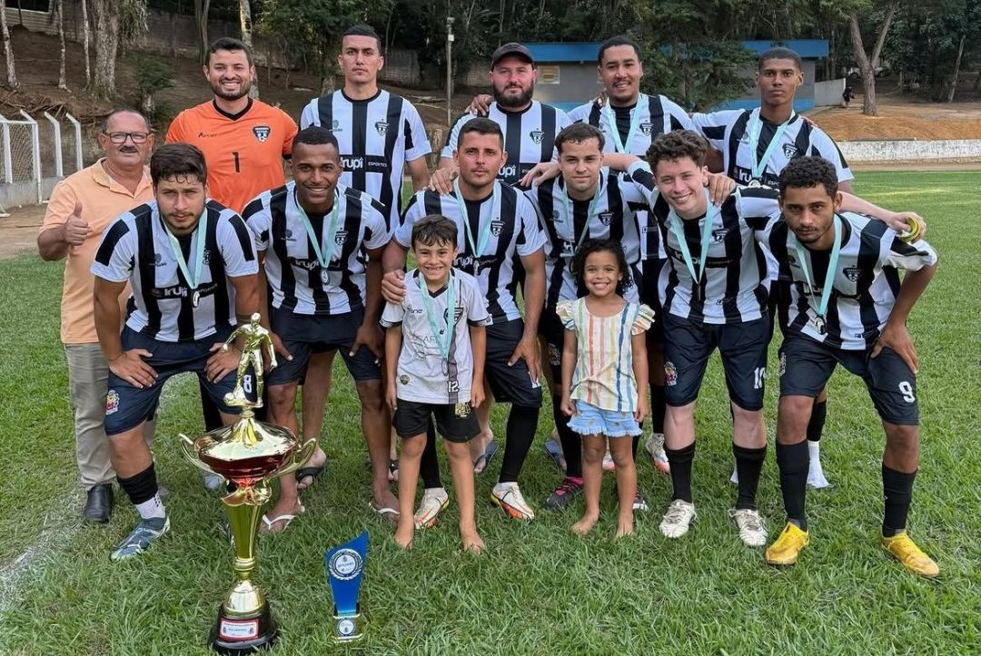 A imagem mostra a equipe do Resenha, vice-campeã do Torneio do Trabalhador, posando para a foto oficial. O time, composto por onze jogadores adultos e duas crianças, está posicionado em pé e agachado em duas fileiras sobre a grama. A maioria dos jogadores veste o uniforme oficial: uma camisa com listras verticais pretas e brancas, calções pretos e meias pretas. Todos os jogadores usam medalhas douradas com fitas azuis claras no pescoço. À frente, no gramado, destaca-se um grande troféu dourado e prateado com uma estatueta de jogador de futebol no topo, sobre uma base preta que contém um adesivo azul e branco. O fundo mostra a lateral do campo com alambrado e, além dele, uma área de mata verde densa sob luz natural do dia.