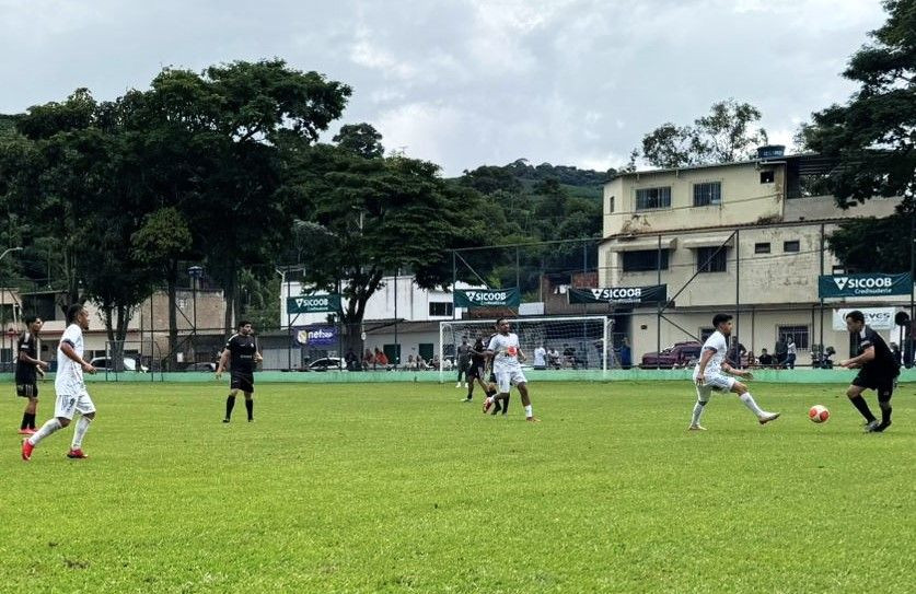 Fotografia de uma partida de futebol de campo durante o dia. Em um gramado verde, jogadores vestindo uniformes brancos e pretos disputam a posse da bola. Ao fundo, árvores altas, casas e faixas de publicidade do Sicoob Credisudeste delimitam o campo. O céu está nublado.