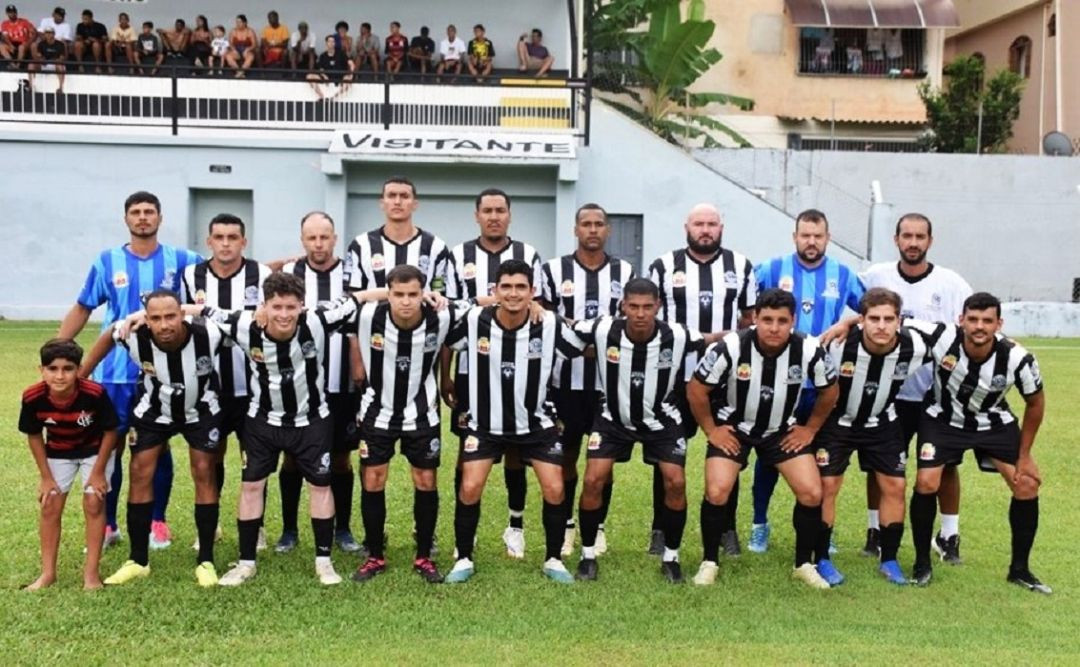 Fotografia oficial da equipe masculina de Irupi reunida no gramado. Os jogadores vestem uniforme com listras verticais em preto e branco. Parte do grupo está em pé e a outra parte agachada. Ao fundo, vê-se a arquibancada com torcedores e a placa indicando o setor 