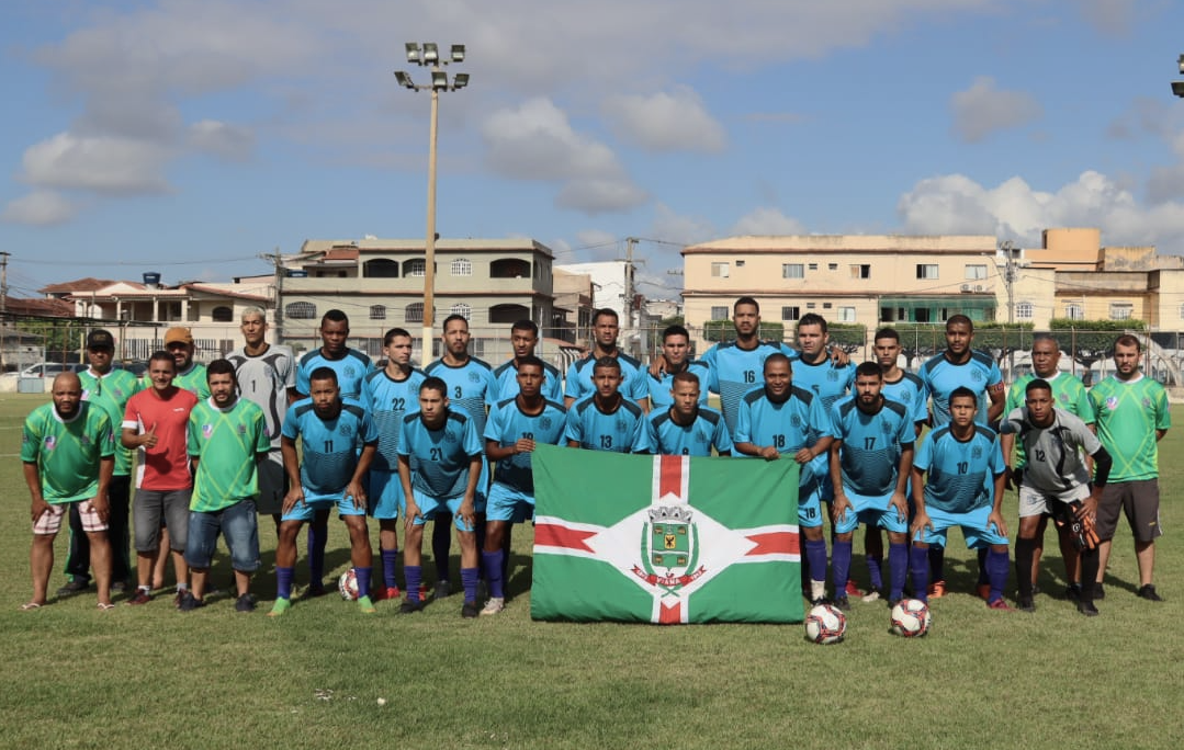 Fotografia oficial da equipe masculina de Viana posicionada no centro do campo. Os jogadores vestem uniformes azul-celeste com detalhes em preto. Dois membros da comissão técnica usam camisas verdes. À frente do grupo, dois jogadores seguram a bandeira oficial do município de Viana. Ao fundo, prédios residenciais e refletores do estádio sob céu com algumas nuvens.