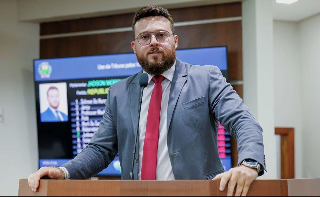 O vereador Jadson Alves Moreno, de barba, óculos e terno cinza, com gravata vermelha, discursando em uma tribuna de madeira. Ao fundo, um painel eletrônico com nomes e informações legislativas.