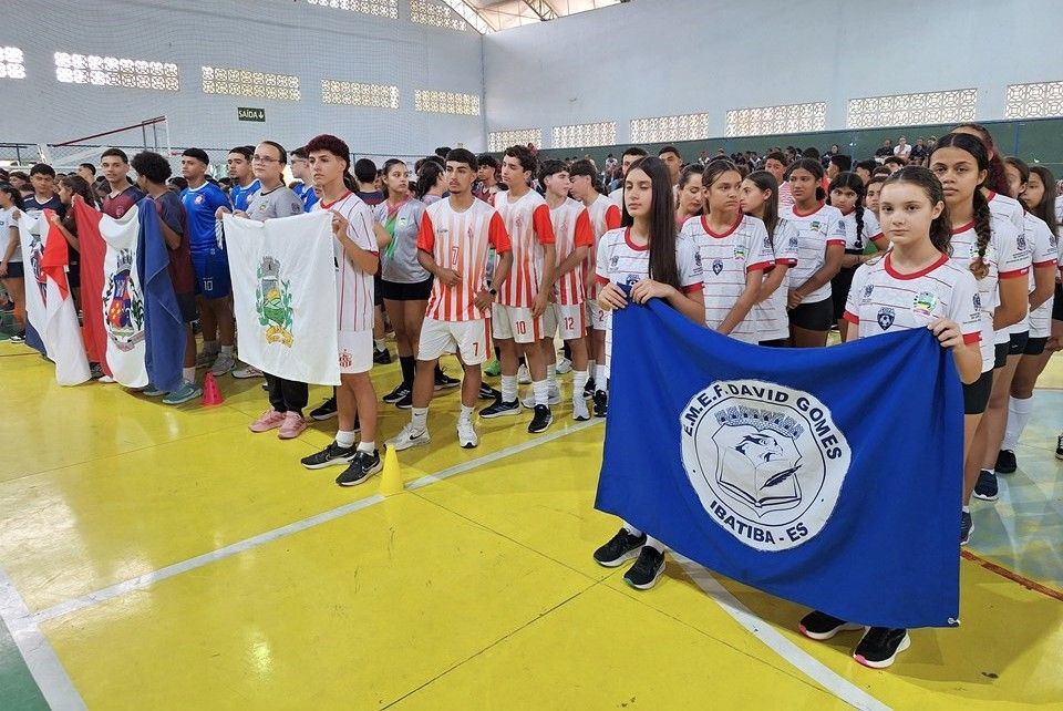 Estudantes uniformizados participam da abertura dos Jogos Escolares em ginásio, organizados em equipes e segurando bandeiras de escolas na regional realizada em Guaçuí, no Espírito Santo.