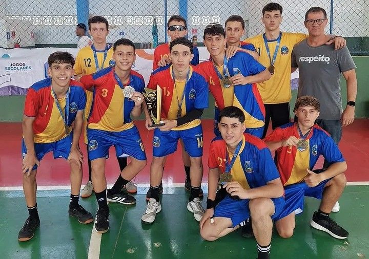 Uma fotografia colorida de um grupo de nove jovens atletas e seu técnico em pé, posando para uma foto de equipe após uma vitória no vôlei. Eles estão dentro de um ginásio poliesportivo com piso verde. Os atletas vestem uniformes de jogo combinando: camisas com divisões em azul, vermelho e amarelo, e shorts azuis, quase todos usando medalhas de ouro ou prata penduradas no pescoço.