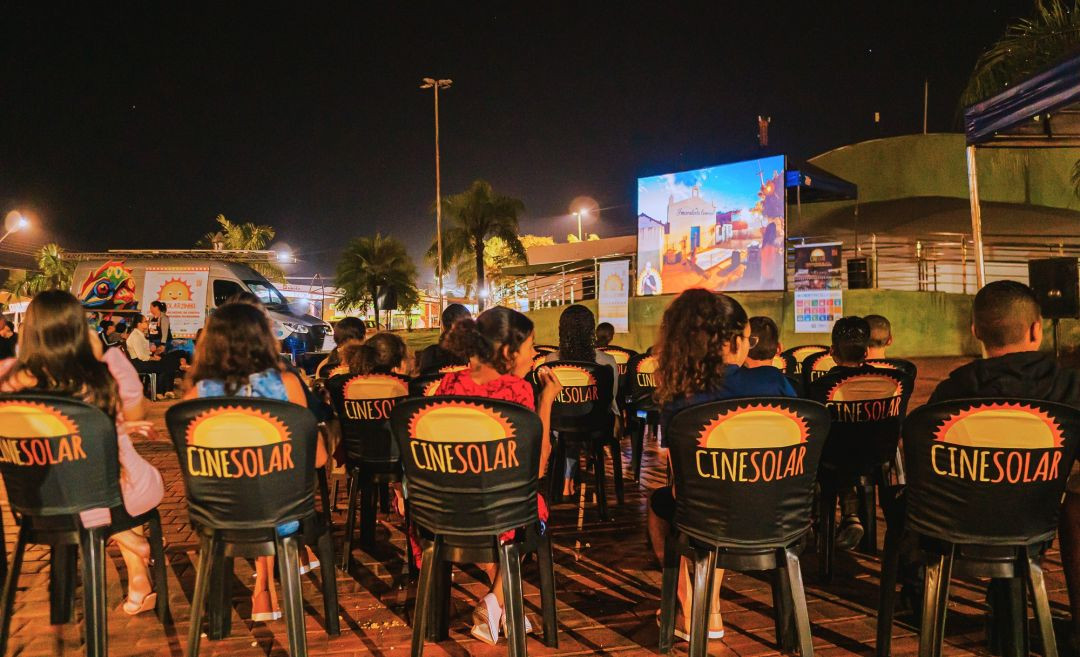 Fotografia noturna de uma plateia ao ar livre assistindo a um filme em um telão. As cadeiras pretas possuem capas com o logotipo 