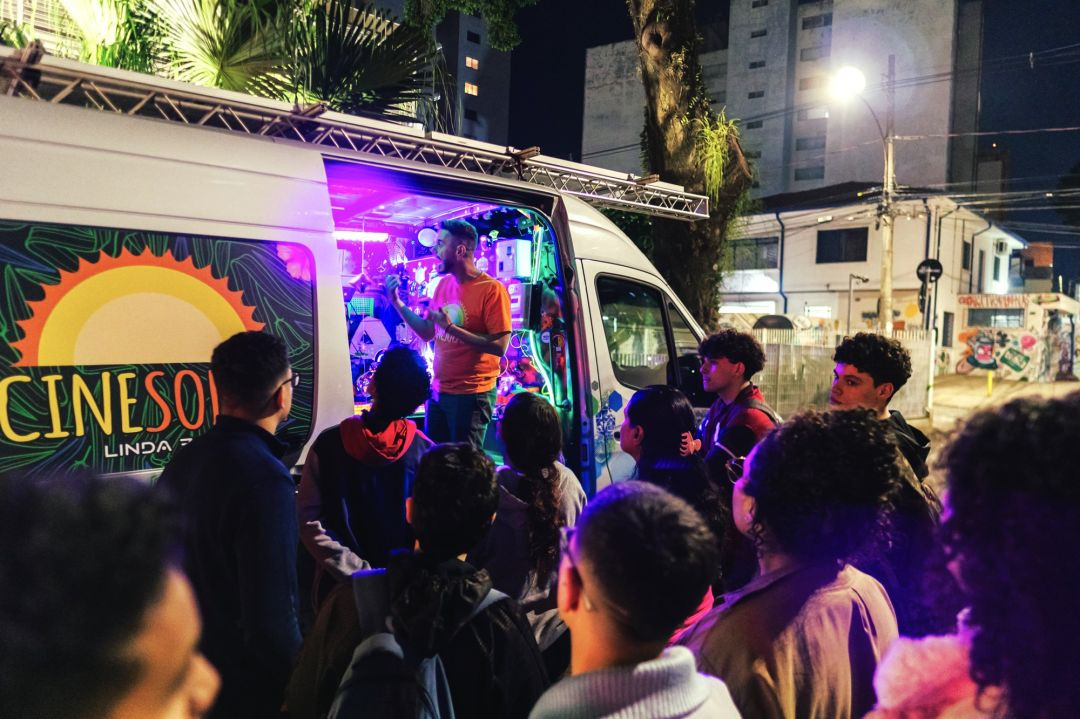 Fotografia noturna mostrando a lateral de um furgão branco adaptado do projeto CineSolar, estacionado em uma rua da cidade. A porta lateral está aberta e revela um interior iluminado por luzes roxas neon e equipamentos eletrônicos. Um homem vestindo uma camiseta laranja com o logo do projeto está de pé dentro do veículo, falando e gesticulando para um grupo de jovens que o escuta atentamente do lado de fora. Na lateral do furgão, lê-se parcialmente a logo 