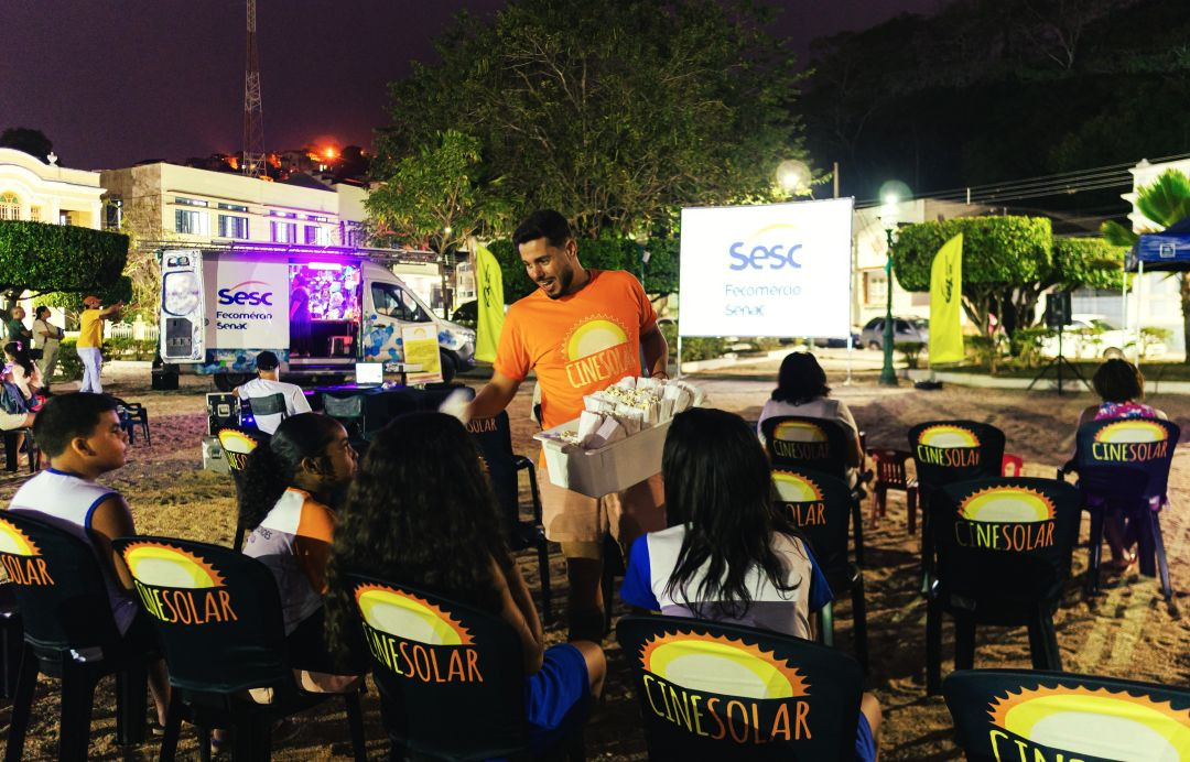 Foto em plano médio capturada durante a noite em uma praça. No centro, um homem com camiseta laranja do CineSolar sorri enquanto segura e oferece uma caixa cheia de saquinhos de pipoca para crianças sentadas em cadeiras de plástico pretas. As cadeiras têm capas pretas com o logo 