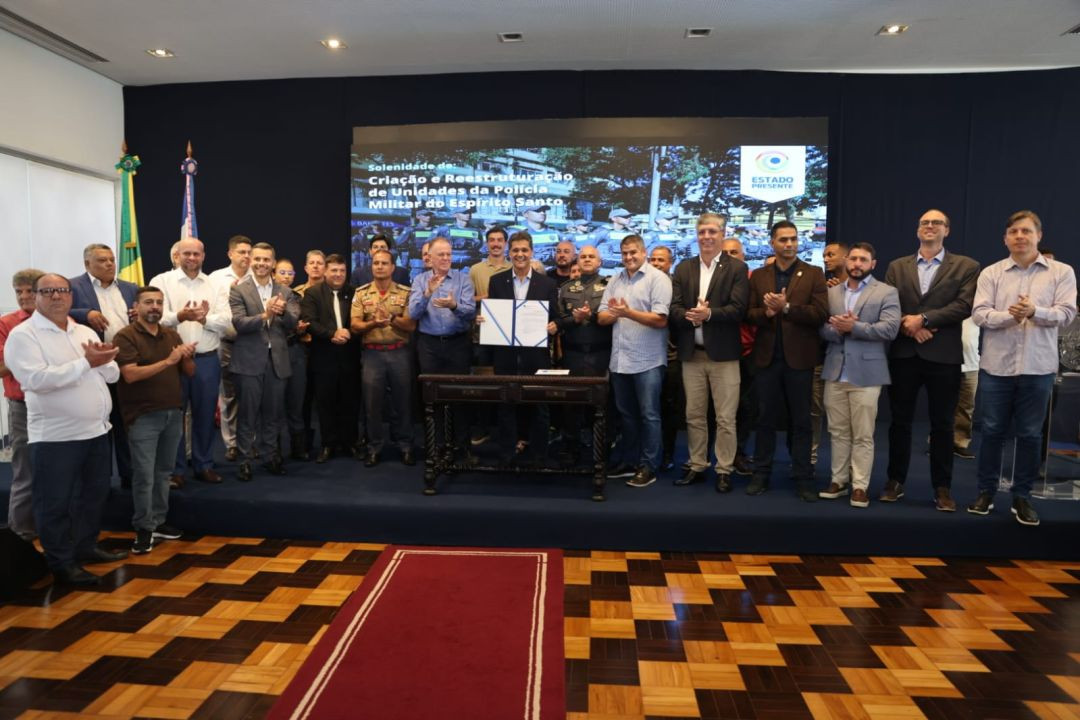 O governador Ricardo Ferraço ao centro, segurando um documento oficial aberto, rodeado por secretários, deputados e oficiais da Polícia Militar e do Corpo de Bombeiros em um palco com tapete azul. Ao fundo, um telão exibe o texto 