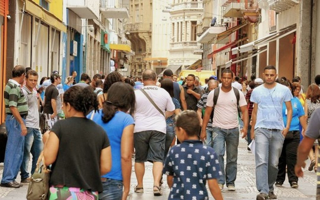 Fotografia em plano médio de uma rua comercial movimentada em um centro urbano brasileiro. Diversas pessoas de diferentes idades e vestimentas caminham em ambos os sentidos sobre um calçadão de pedras portuguesas. Ao fundo, prédios históricos e fachadas de lojas com toldos coloridos compõem o cenário sob luz natural do dia.