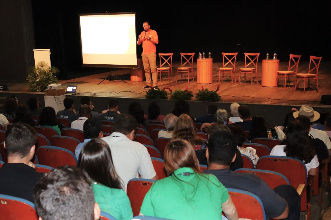 Homem com camisa laranja discursa em um palco de teatro ao lado de uma tela de projeção branca. Ao fundo, cadeiras de madeira e mesas de apoio organizadas para um painel. Auditório com público sentado.