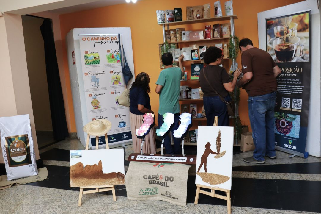 Visitantes observam uma estante com diversos pacotes de café de diferentes marcas. Ao lado, um painel explicativo intitulado 