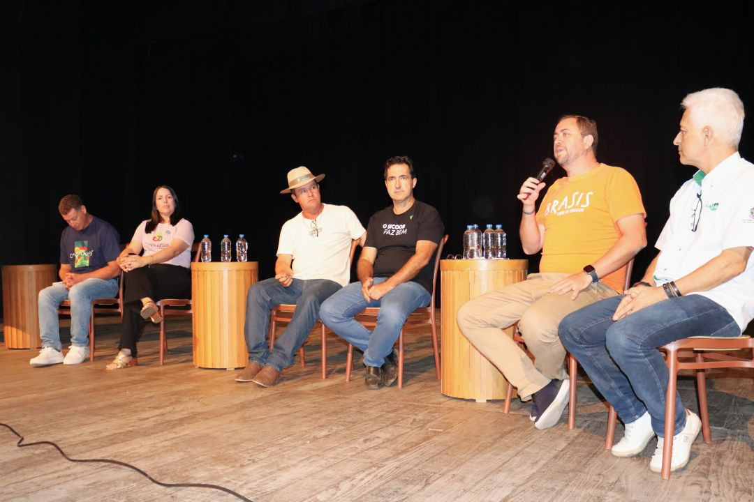 Seis pessoas sentadas em cadeiras de madeira no palco durante um debate. O homem ao centro, com camisa laranja, fala ao microfone. Há garrafas de água sobre mesas laterais de madeira.