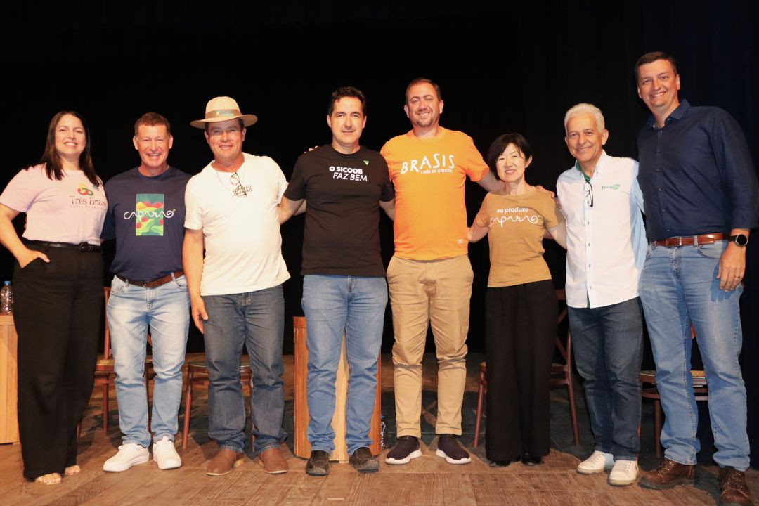 Oito pessoas (homens e mulheres) posadas lado a lado no palco para uma foto oficial. Elas sorriem e vestem roupas casuais e camisas de instituições parceiras.