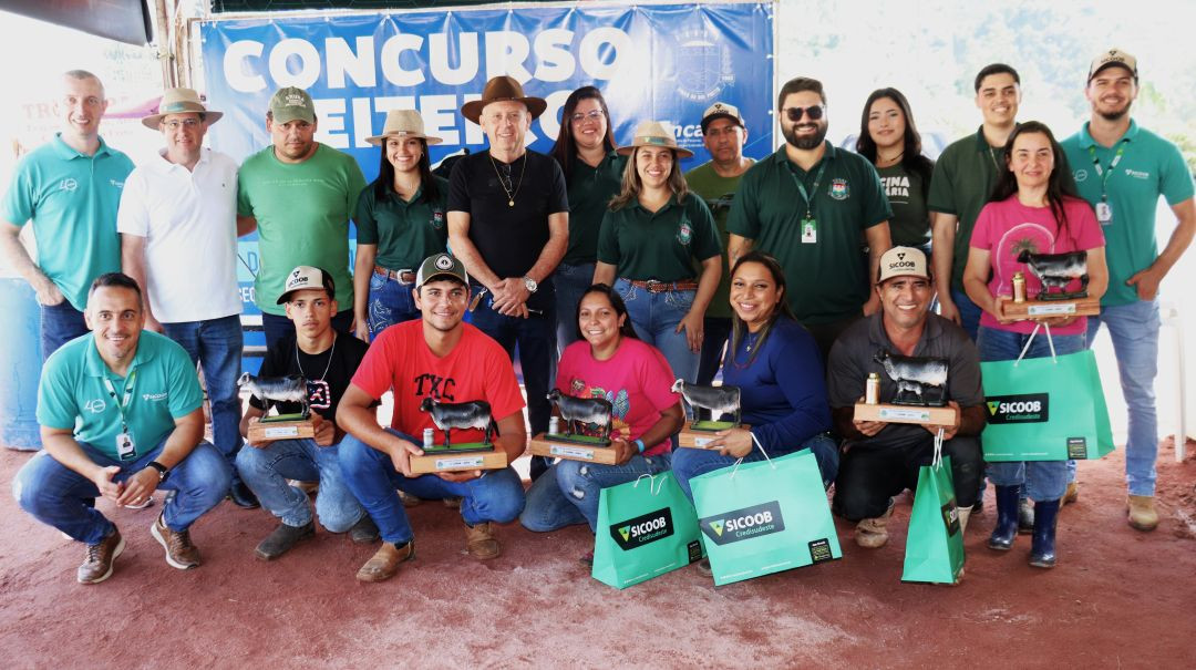 Foto oficial de um grupo de cerca de 20 pessoas posando para a câmera. Alguns homens agachados à frente seguram troféus de vacas leiteiras e sacolas de um patrocinador (Sicoob). Ao fundo, um banner azul com os dizeres 