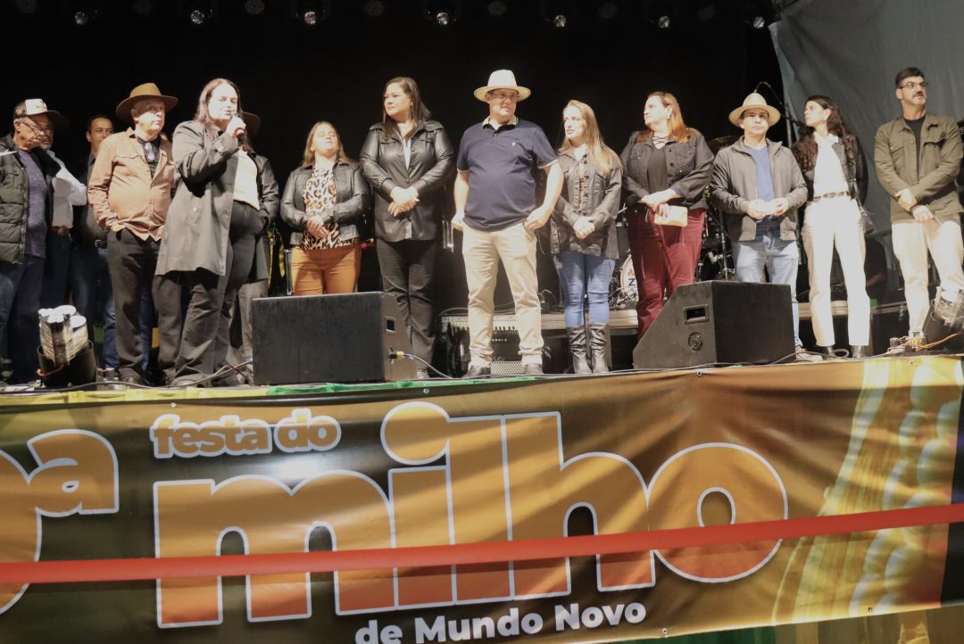 Grupo de autoridades e representantes da comunidade perfilados em cima do palco durante a abertura oficial. À frente, um banner horizontal com a logomarca 