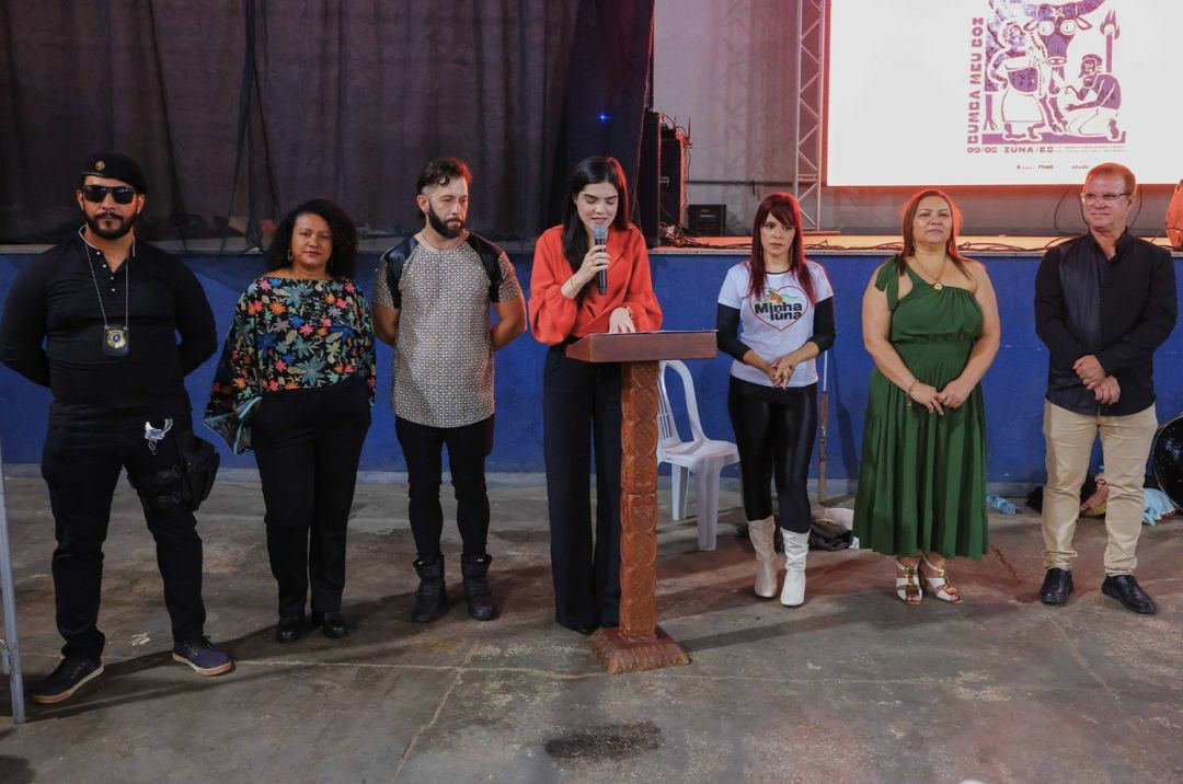Autoridades e organizadores posicionados em frente ao palco. Uma mulher ao centro fala ao microfone em um púlpito de madeira. Ao fundo, um telão exibe a identidade visual do evento com o tema 