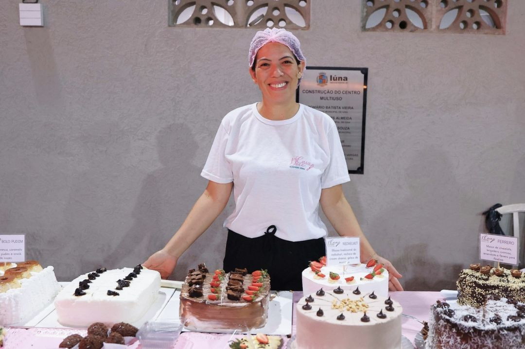 Mulher sorridente atrás de uma mesa de doces na praça de alimentação. Sobre a mesa, diversos bolos decorados, incluindo sabores como red velvet, chocolate e bolo pudim, com etiquetas de identificação.