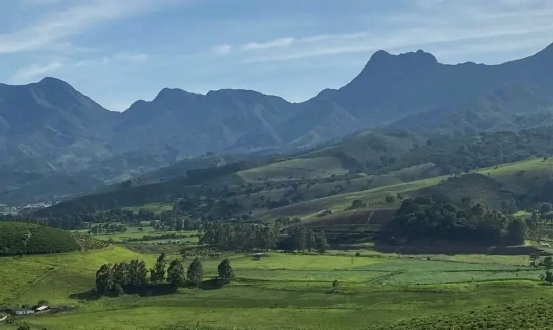 Vista panorâmica da região de Pedra Menina, em Dores do Rio Preto. A imagem exibe um vale com vastas áreas de pastagens e plantações verdes em diferentes tons. Ao fundo, destaca-se a imponente silhueta das montanhas da Serra do Caparaó sob um céu com nuvens esparsas.