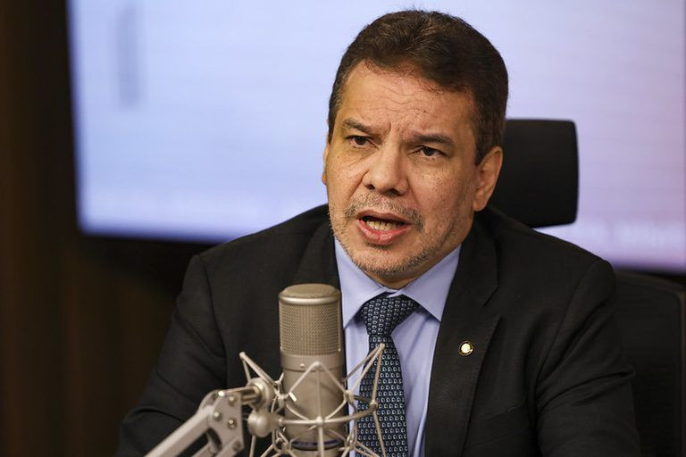Fotografia em plano médio do Ministro da Justiça e Segurança Pública, Wellington Lima. Ele é um homem de pele parda, cabelos escuros e curtos, veste terno escuro, camisa azul clara e gravata azul marinho. Ele está falando em um microfone de estúdio de rádio, com semblante sério e focado. Ao fundo, uma tela de monitor desfocada.