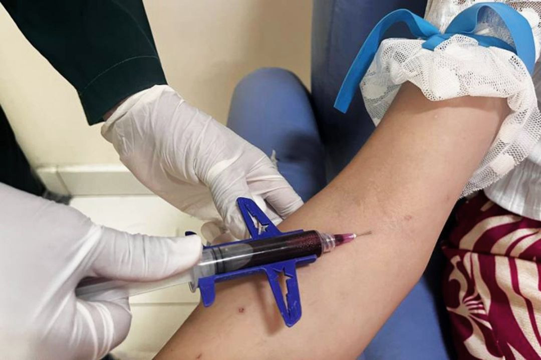 Foto em plano detalhe de um profissional de saúde, usando luvas brancas, realizando uma coleta de sangue no braço de um paciente com uma seringa acoplada a um dispositivo azul.