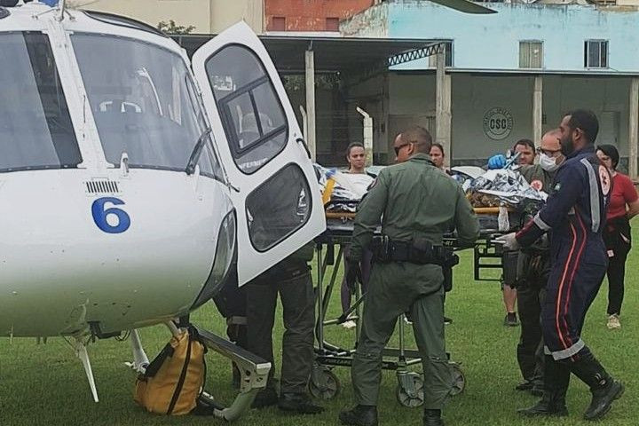 Foto mostra profissionais do Samu e do Notaer colocando o paciente vítima de AVC, em Muniz Freire, no helicóptero, em um campo gramado de futebol, com outras pessoas observando.