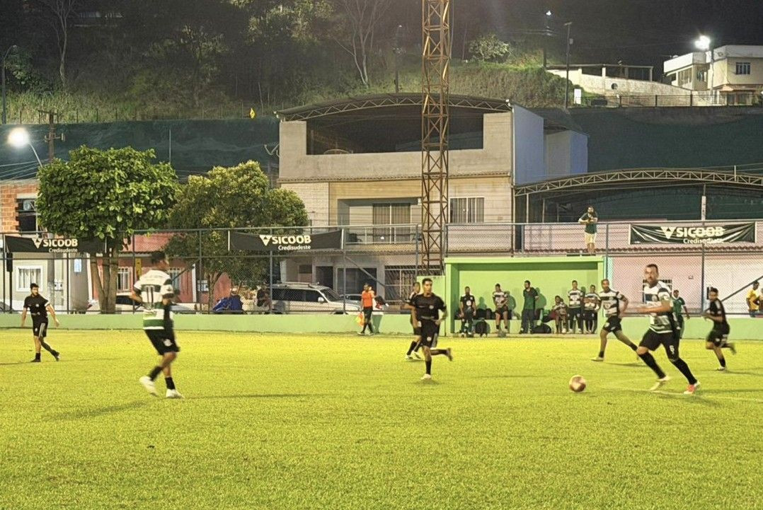 Registro em movimento de uma partida de futebol noturna, sob a luz dos refletores. Jogadores de duas equipes — uma vestindo uniforme listrado em verde e branco e a outra vestindo uniforme preto — disputam a posse da bola no meio de campo. Ao fundo, vê-se a estrutura metálica de iluminação, arquibancadas com faixas do Sicoob e residências na encosta ao fundo.