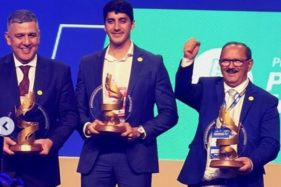 Imagem horizontal em plano médio mostrando três homens no palco de uma premiação, com um painel azul e verde ao fundo. Todos vestem trajes formais e seguram troféus dourados idênticos em formato de espiral. O homem da direita, o prefeito Paulino Lourenço, veste terno azul, usa óculos e comemora erguendo o punho direito fechado.