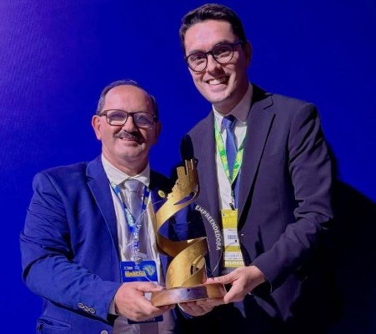 Fotografia em plano médio de dois homens sorrindo em um evento formal, com fundo azul texturizado. À esquerda, o prefeito Paulino Lourenço usa óculos, bigode e terno azul com crachá do evento. À direita, um homem mais jovem de óculos e terno escuro, o vice-prefeito Abercílio Machado. Juntos, eles seguram um troféu dourado em formato espiralado e vertical, com base de madeira.