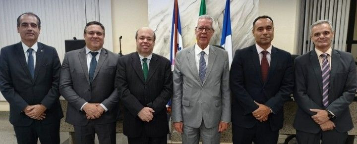 Foto oficial de plano médio mostrando seis homens alinhados lado a lado em um ambiente institucional com detalhes em mármore claro ao fundo. Todos vestem paletó e gravata em tons escuros e cinza. Da esquerda para a direita, homens de meia-idade posam formalmente com as mãos cruzadas à frente do corpo. Ao centro-direito, um homem grisalho de óculos veste terno cinza claro.