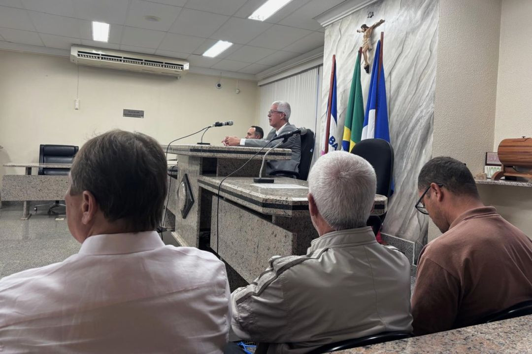 Visão lateral de dentro do plenário do júri. Em primeiro plano, vê-se a parte de trás da cabeça e ombros de três homens sentados assistindo à sessão. Ao fundo, em uma mesa elevada de mármore sob um crucifixo fixado na parede e três bandeiras oficiais, o corregedor-geral, um homem grisalho de terno cinza, fala ao microfone.