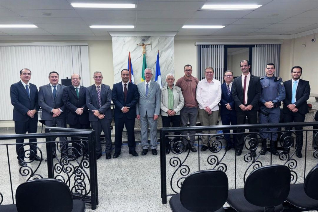 Foto grupal em plano aberto registrando as autoridades presentes no Auditório do Tribunal do Júri de Iúna. Catorze homens estão posicionados de pé em linha horizontal atrás de uma grade de proteção preta. Entre eles, há civis em trajes formais (terno e gravata), homens em trajes casuais e um policial militar fardado. Ao fundo, a parede de mármore abriga as bandeiras do Brasil, do Espírito Santo e o crucifixo.