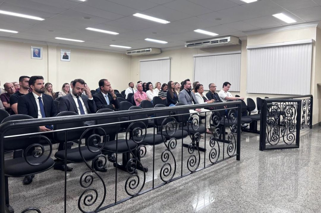 Foto ampla mostrando o público assistindo à solenidade no auditório. Diversas fileiras de cadeiras pretas estofadas estão ocupadas por homens e mulheres trajando roupas formais e casuais. A sala possui piso de granito polido cinza, luminárias de LED embutidas no teto, aparelhos de ar-condicionado nas paredes e janelas fechadas com persianas brancas.