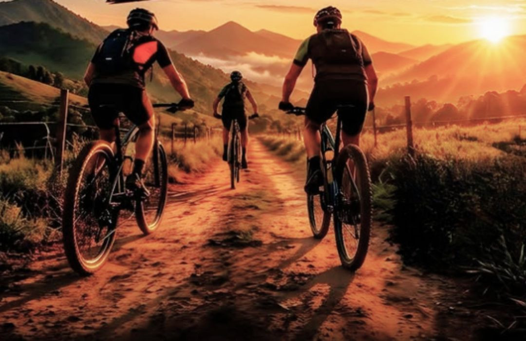Três ciclistas são vistos de costas pedalando por uma estrada de terra batida e poeira durante o amanhecer. O sol nasce forte no horizonte, entre montanhas cobertas por neblina, iluminando o caminho com tons quentes de laranja e dourado. Os ciclistas vestem roupas esportivas e mochilas de hidratação.