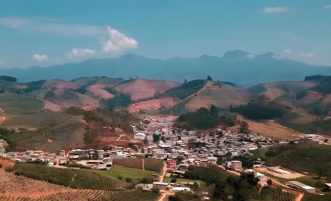 Vista aérea panorâmica da cidade de Irupi, localizada na região do Caparaó, no Espírito Santo. O aglomerado urbano, composto por casas e edifícios de poucos andares de cores predominantemente claras, estende-se ao longo de um vale. O entorno é caracterizado por colinas onduladas com plantações de café dispostas em linhas paralelas e áreas de solo exposto em tons terrosos. Ao fundo, uma cadeia de montanhas imponentes sob um céu azul claro com poucas nuvens define a linha do horizonte.