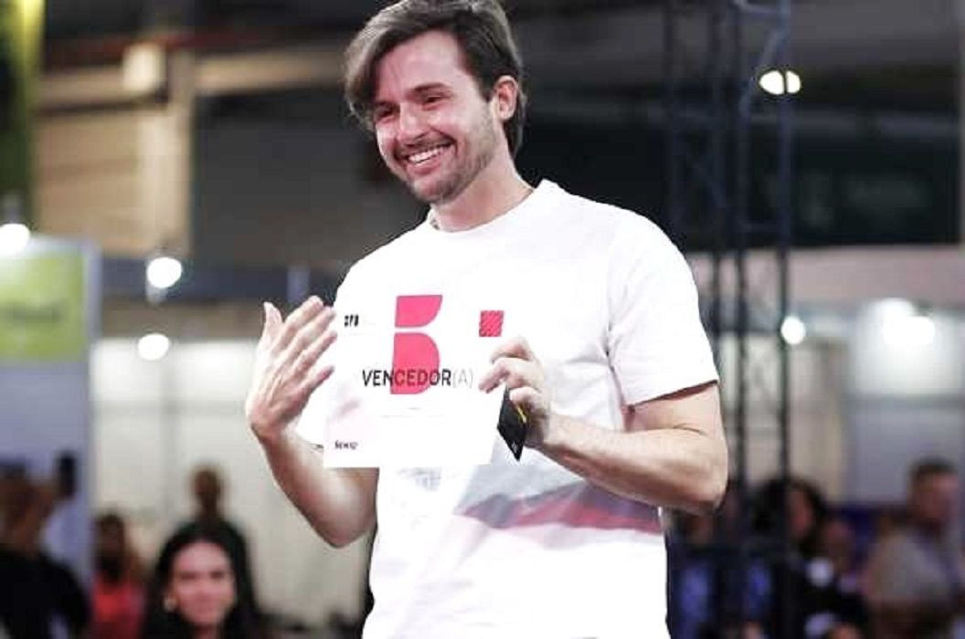Fotografia em plano médio focada no estilista Maurício Mazzon Magnoni no palco do evento. Ele está de pé, sorrindo, com o corpo ligeiramente voltado para a esquerda. Ele usa uma camiseta branca com uma estampa vermelha e segura com as duas mãos um certificado em papel branco onde se lê a palavra 