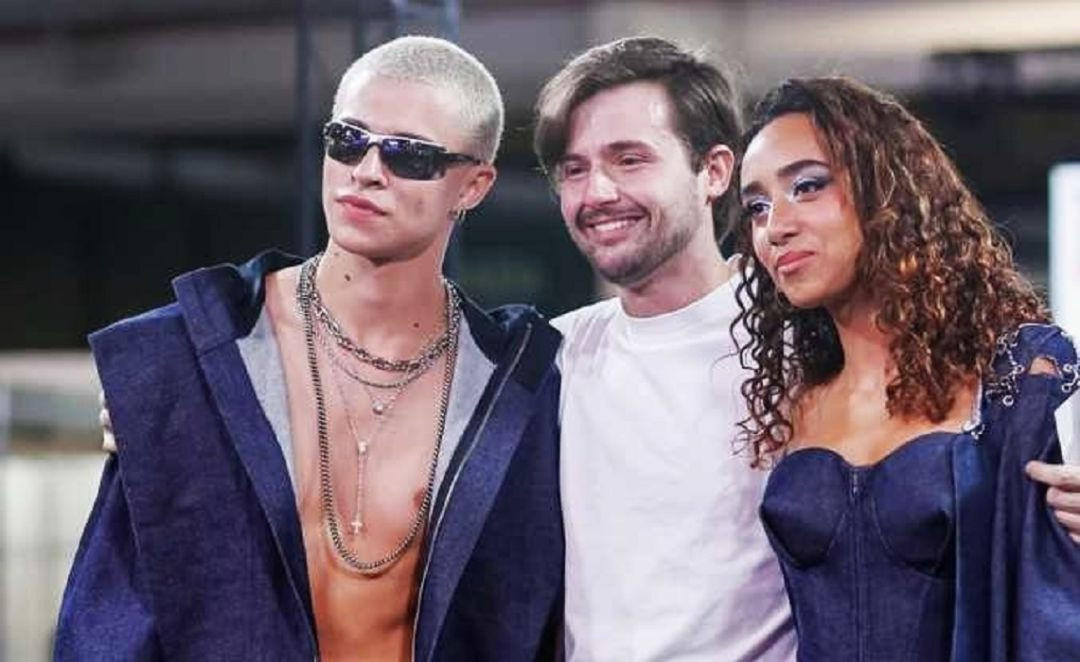 Fotografia colorida de três pessoas em plano médio, do peito para cima, em um evento de moda. À esquerda, um jovem modelo com cabelos loiros raspados e óculos de sol escuros usa colares de corrente e um casaco jeans aberto. No centro, o estilista Maurício Mazzon Magnoni, um homem branco de cabelos castanhos e barba aparada, sorri vestindo uma camiseta branca. À direita, uma modelo negra com cabelos cacheados e maquiagem azul nos olhos veste um espartilho jeans.