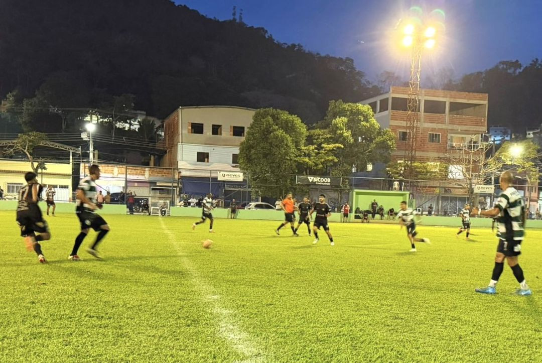 Fotografia noturna de uma partida de futebol de campo em andamento no Estádio Municipal de Dores do Rio Preto. O gramado verde está bem iluminado por uma alta torre de refletores acesa no lado direito, sob um céu de fim de tarde em tom azul-escuro. Jogadores correm pelo campo; parte do time veste uniforme listrado em verde e branco com calções pretos, e outros usam uniforme totalmente escuro. Ao fundo, atrás do alambrado, avistam-se árvores, construções urbanas e faixas comerciais com as marcas Sicoob Credisudeste.