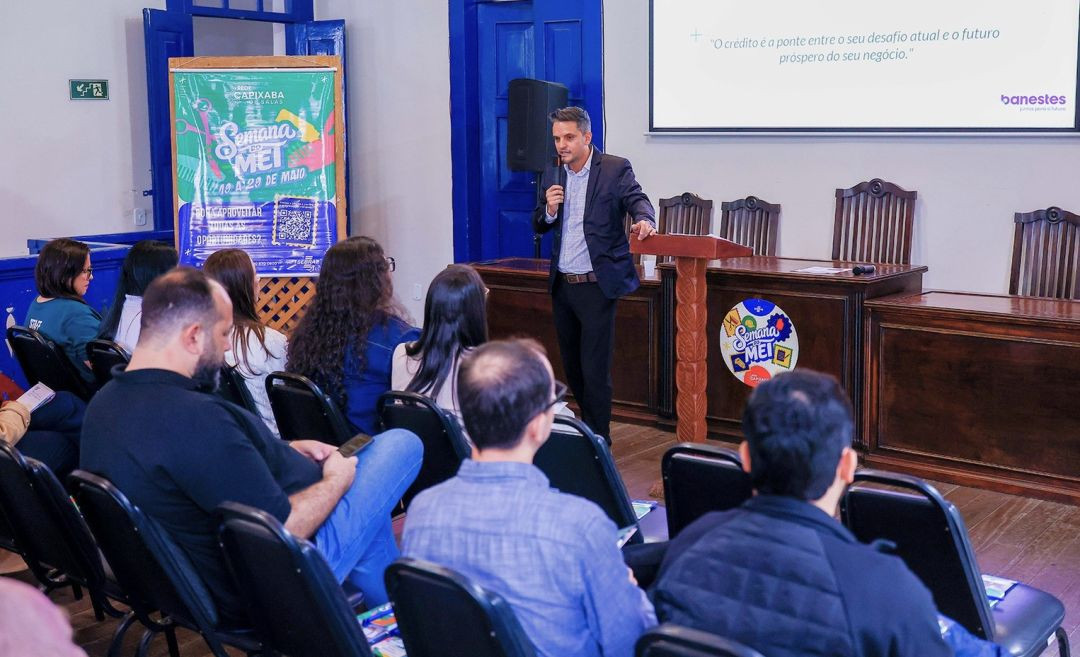 Fotografia em plano médio da palestra na Casa da Cultura de Iúna. À direita, um homem de terno escuro e camisa clara fala ao microfone atrás de um púlpito de madeira. Ao fundo, uma tela de projeção exibe a frase promocional do Banestes sobre crédito. À esquerda, o público assiste sentado em cadeiras pretas. Em destaque na parede, um banner colorido traz a inscrição 