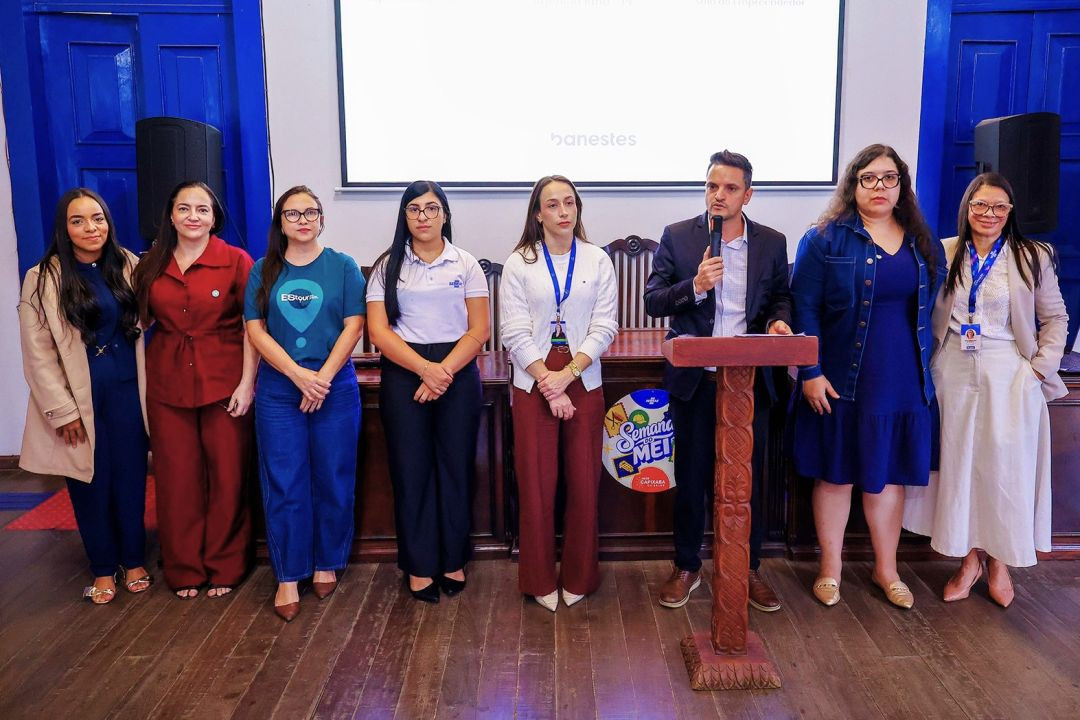 Foto oficial com o grupo de autoridades e palestrante alinhados lado a lado no palco de madeira da Casa da Cultura. Da esquerda para a direita, estão sete mulheres e um homem (o palestrante, que segura o microfone). Eles vestem trajes formais e casuais. Ao fundo, a tela branca de projeção e portas azuis de madeira compõem o cenário.