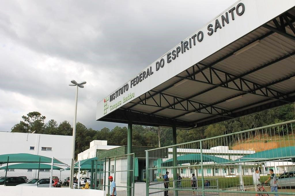 Fotografia da fachada externa do Instituto Federal do Espírito Santo (Ifes) Campus Ibatiba sob céu nublado. Em destaque, uma estrutura de cobertura metálica preta sustenta a identificação visual da instituição com letras em relevo onde se lê 